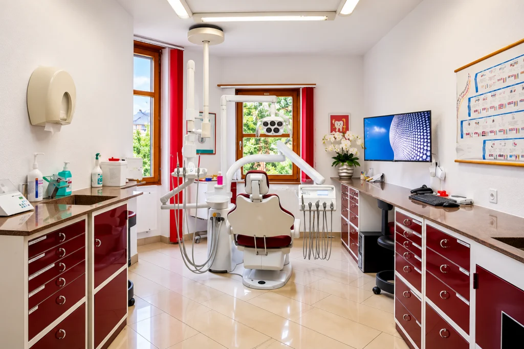 Behandlungszimmer mit dunkelroten Schränken, Behandlungsstuhl und Fenster ins Grüne